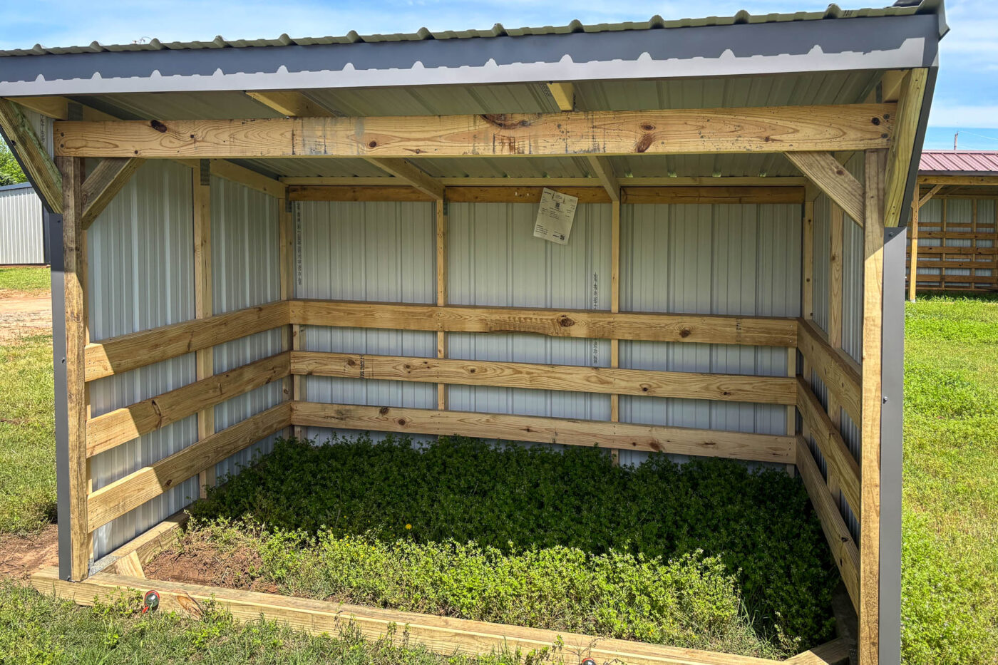 Three-sided wooden loafing shed with partial back wall and full side walls, sloped metal roof, and open front on green grass.
