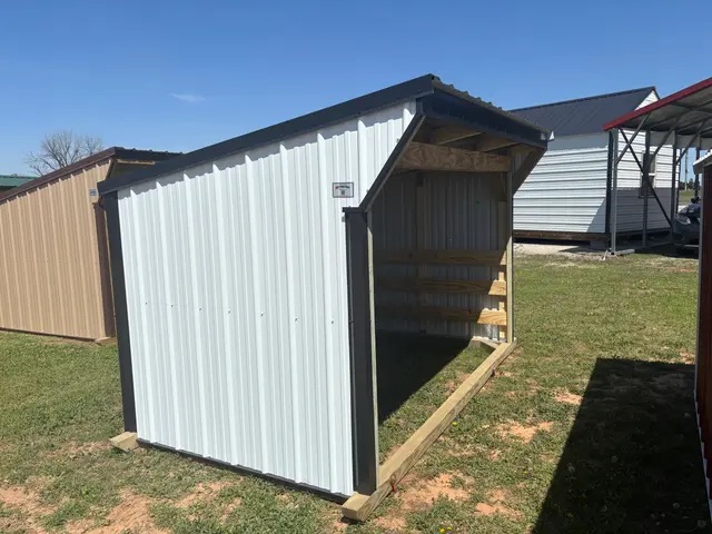 White metal loafing shed with sloped roof and partially enclosed wood interior, standing on grass.