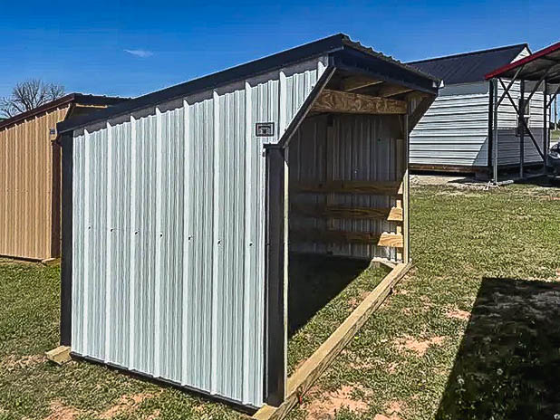 Gray metal loafing shed with sloped roof and open front, supported by wooden framing, on grass.