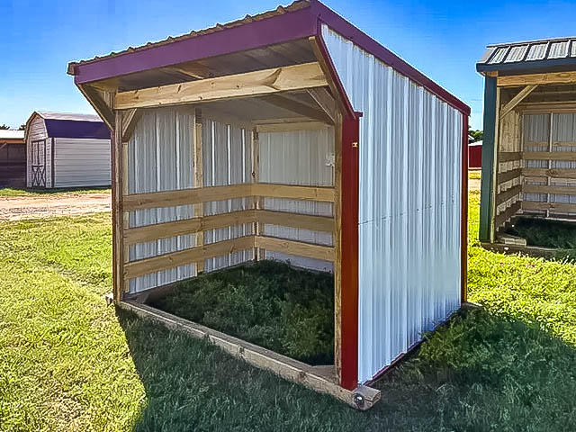 Red and white metal loafing shed with wooden interior framing, three-sided coverage, and sloped roof over green grass.