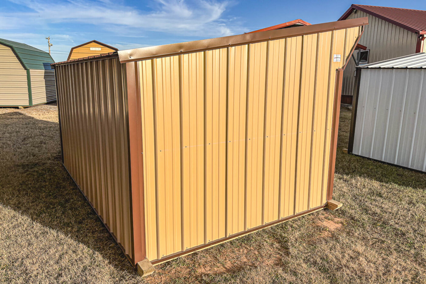 Beige metal loafing shed with red trim and a curved gable-style roof, set on gravel in outdoor lot.