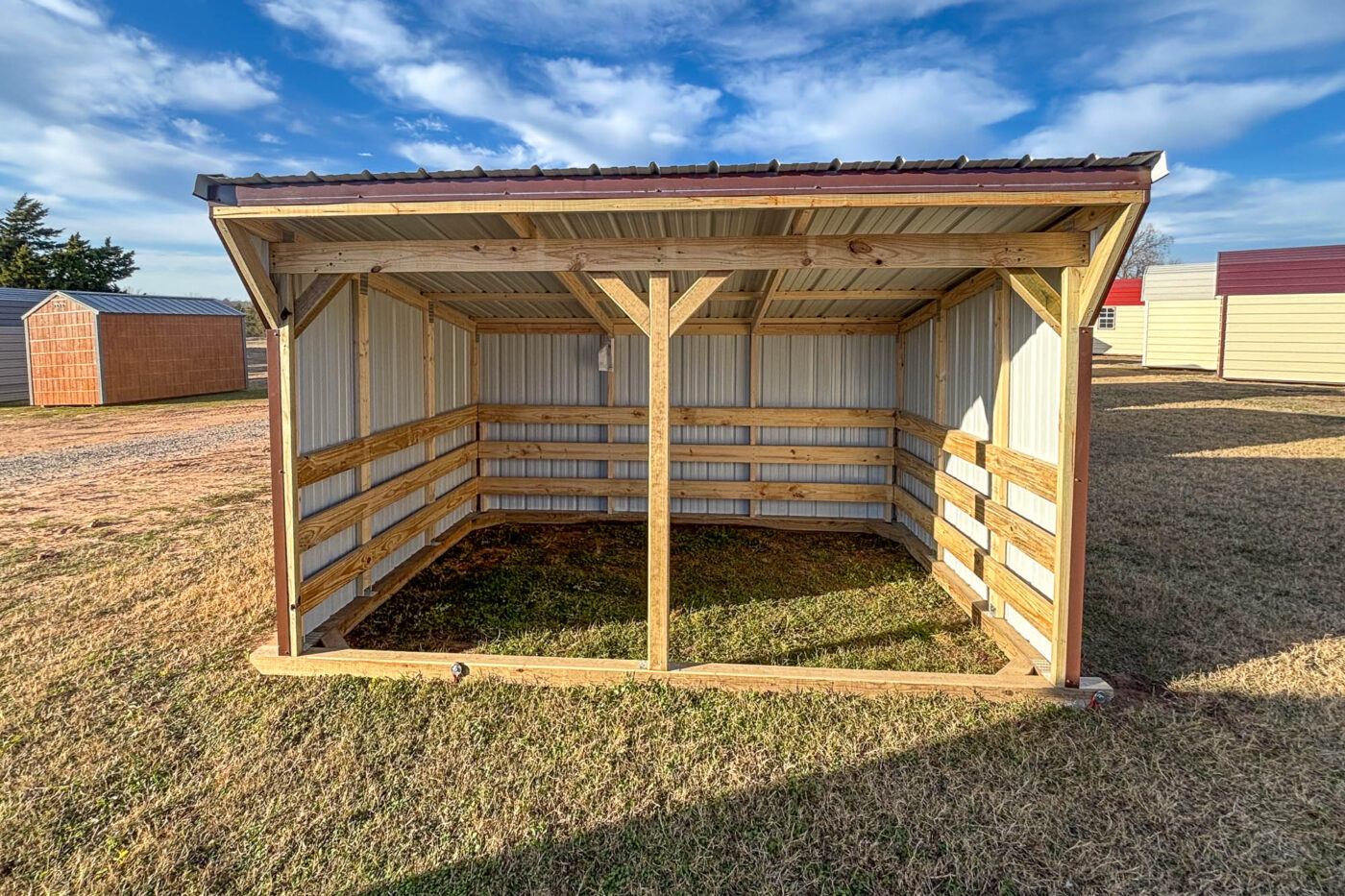 Wooden loafing shed with open front, sloped metal roof, and exposed interior framing, standing on grassy ground under a blue sky.
